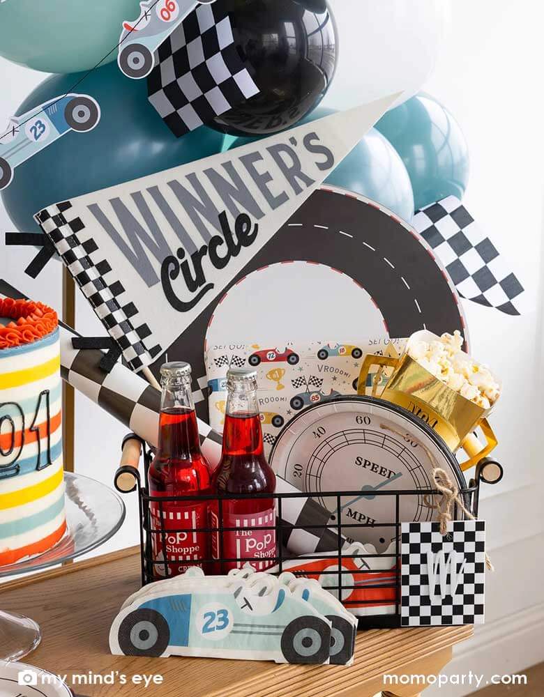 A party occasion bin filled with race car themed party supplies from Momopartys including race track placemats, speedometer shaped paper plates, checkered table runner, race car pattern square plates, race car shaped napkins, and a felt "Winner's Circle" party pennant. Around the basket is a colorful buttercream cake and some party balloons and checkered flags decorated behind the party table. Making this a perfect inspo for kid's race car themed celebrations.