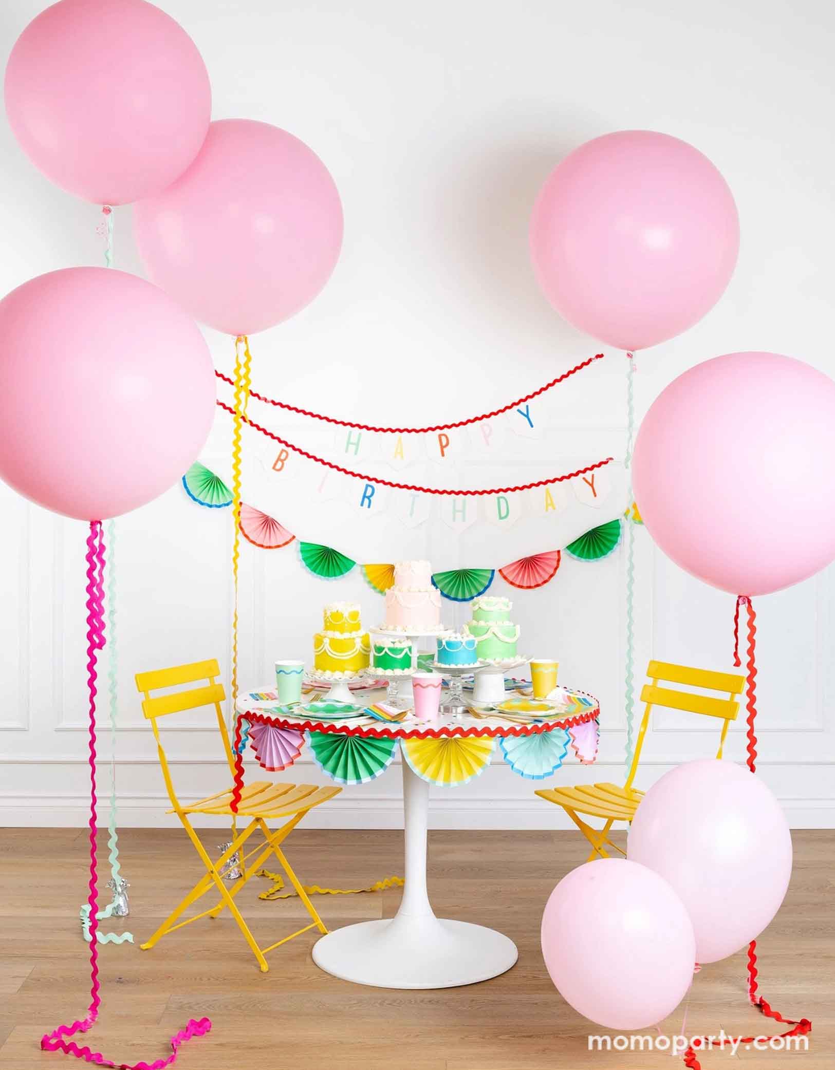 A cheerful and colorful birthday party set up featuring various festive birthday party supplies from Momopartys including rainbow colored tablewares of plates, cups, napkins on the table along with multiple pastel colored birthday cakes. The table is decorated with colorful bunting garland around and in the back of the wall hung a Happy Birthday felt garland and the colorful bunting banner. On the sides are some jumbo pink balloons floating above, making this a whimsical set up for kid's birthday bash!