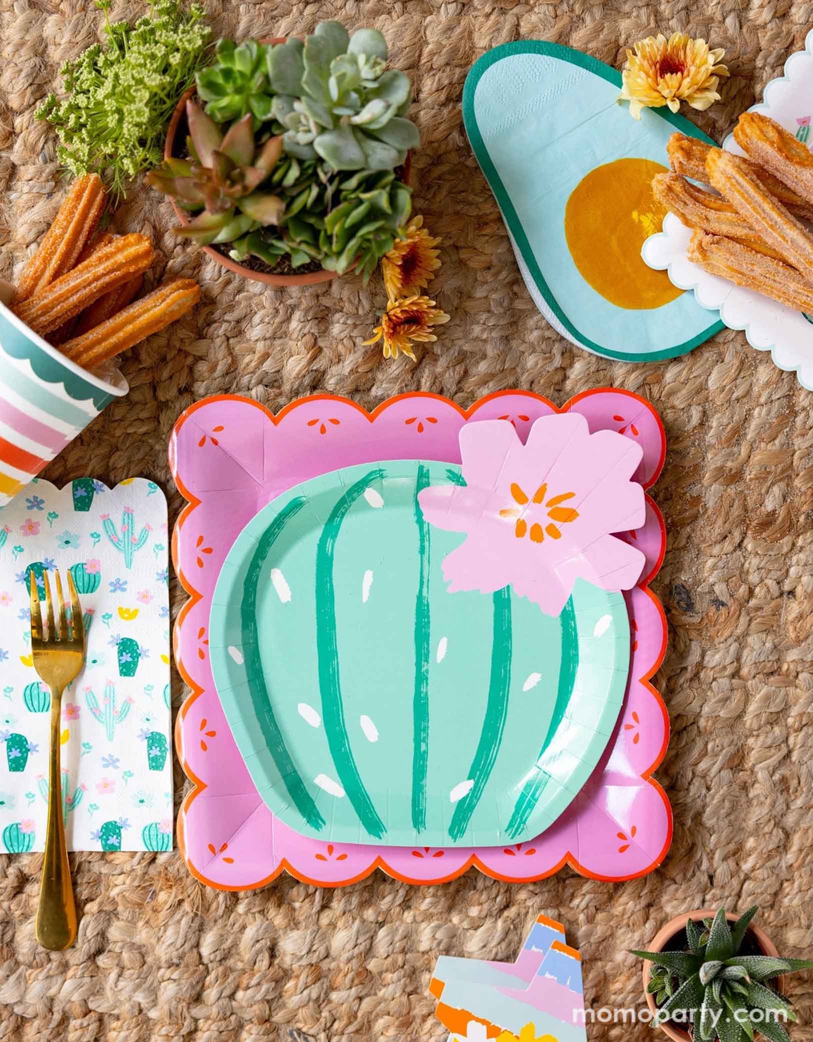 A overhead shot of a fun and playful fiesta themed party tablescape featuring Momopartys's fiesta themed party supplies including the pink Fiesta Papel paper plate, the cactus shaped plate, avocado shaped napkins, and the fiesta rainbow colored striped party cup filled with churros. On the table there are mini pots of cactus and succulent plants, making this a fun table setting idea for a playful yet modern fiesta themed celebration - perfect for girl's "first fiesta" themed first birthday party!