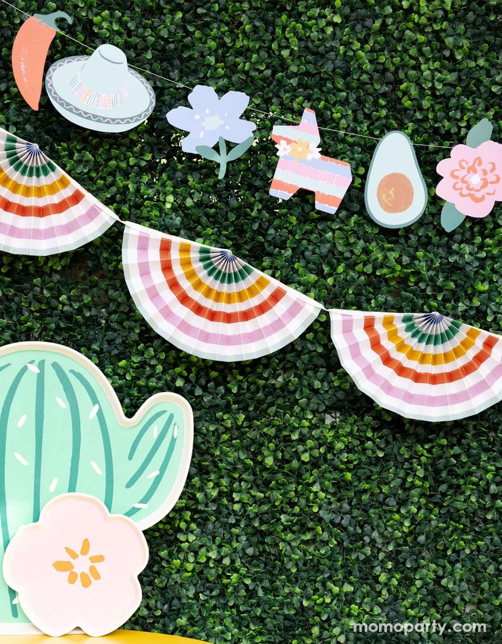 A festive Mexican themed celebration set up featuring Momopartys's Fiesta party supplies including the fiesta icon banner and the rainbow colored bunting garland on the faux leaf wall, along with the cactus shaped serving tray, this makes a simple yet modern look for a fun and playful fiesta celebration!