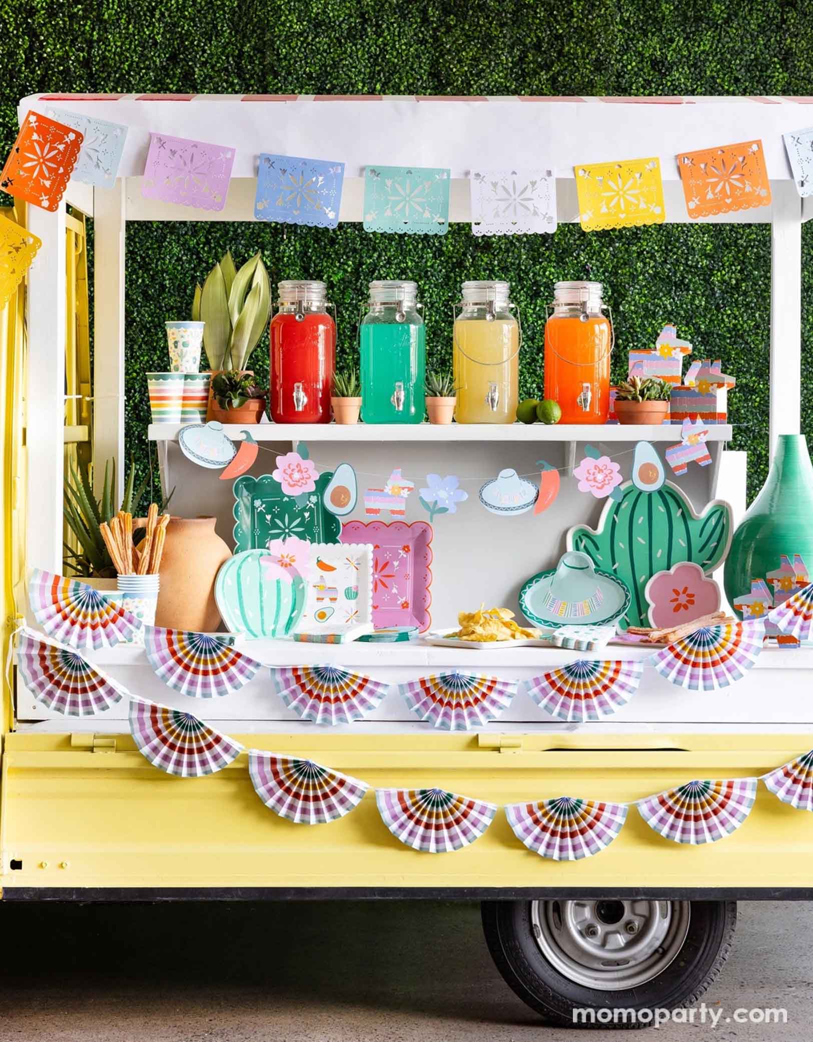 A cheerful yellow Mexican inspired truck is decorated with Momopartys's colorful fiesta party supplies including the Fiesta Papel Picado Banner, bunting banner hung across the truck. In the middle there a various fiesta party tablewares in display including cactus shaped plates, sombrero hat shaped plates, fiesta icon dinner plates, fringe napkins, striped party cups and pinata shaped treat boxes. On the truck there are also colorful drinks in glass pitchers ready to be served for the party guests.