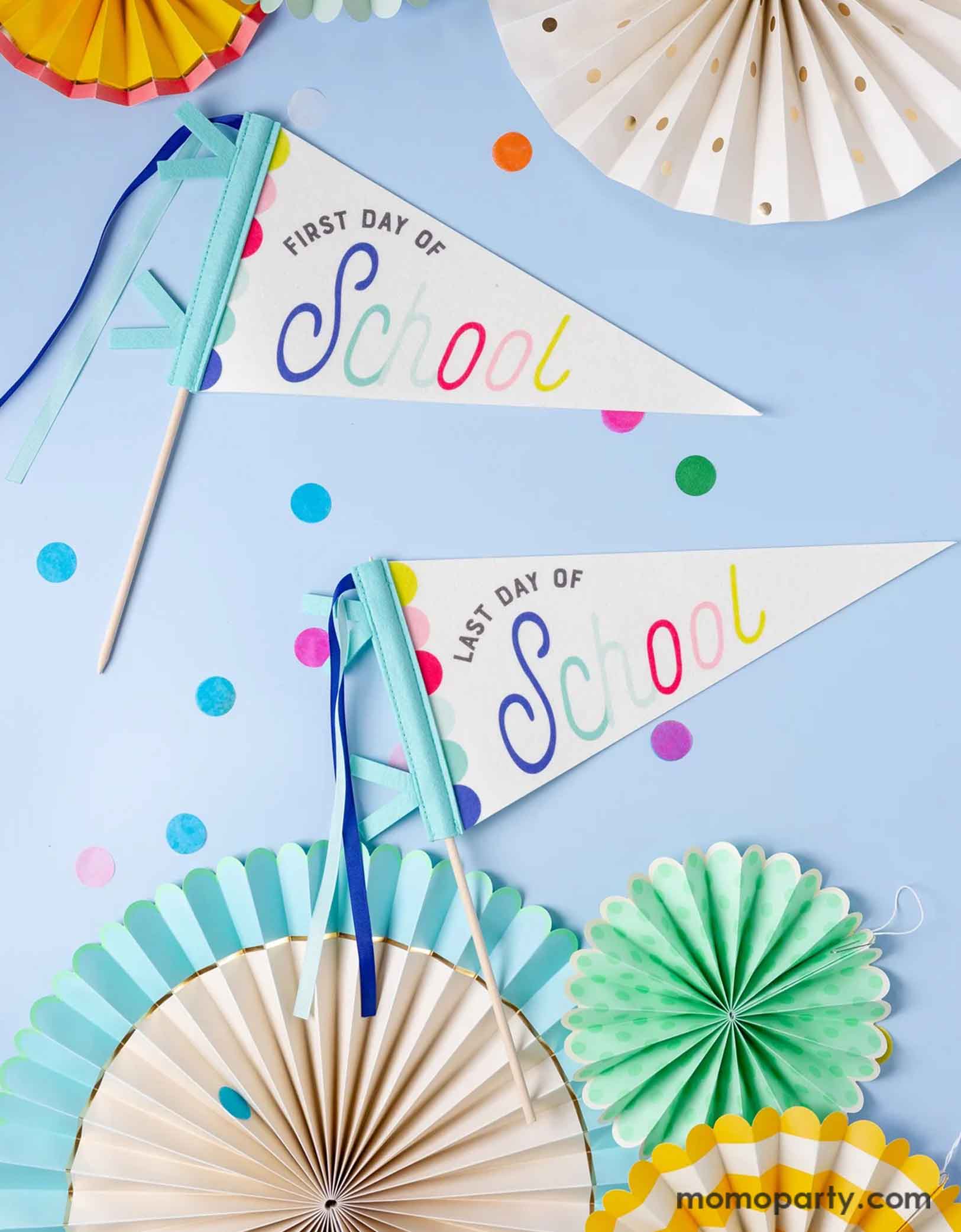 An overhead shot of Momopartys's 14" First Day of School/Last Day of School Pennant Banner Set by My Mind's Eye on a blue background. Around the pennants are colorful paper fans and round confetti.  This playful set features pennants for the first and last day of school, adding a unique touch to your classroom or home decor. Get ready to celebrate and capture those special moments with these festive and fun banners.