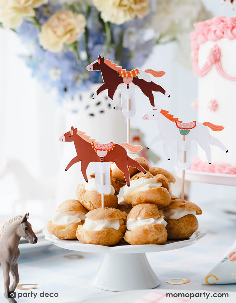 A beautiful horse themed party table featuring some delicious cream puffs stacked topped with Momopartys's floral pony cupcake toppers in 3 different colors and designs. On the table there are some gold horseshoe shaped confetti, a pink and white buttercream cake and a beautiful bouquet in lilac and cream colors, with the horse figurine as the centerpiece, this gives a charming and darling idea for kid's pony or horse themed birthday party.