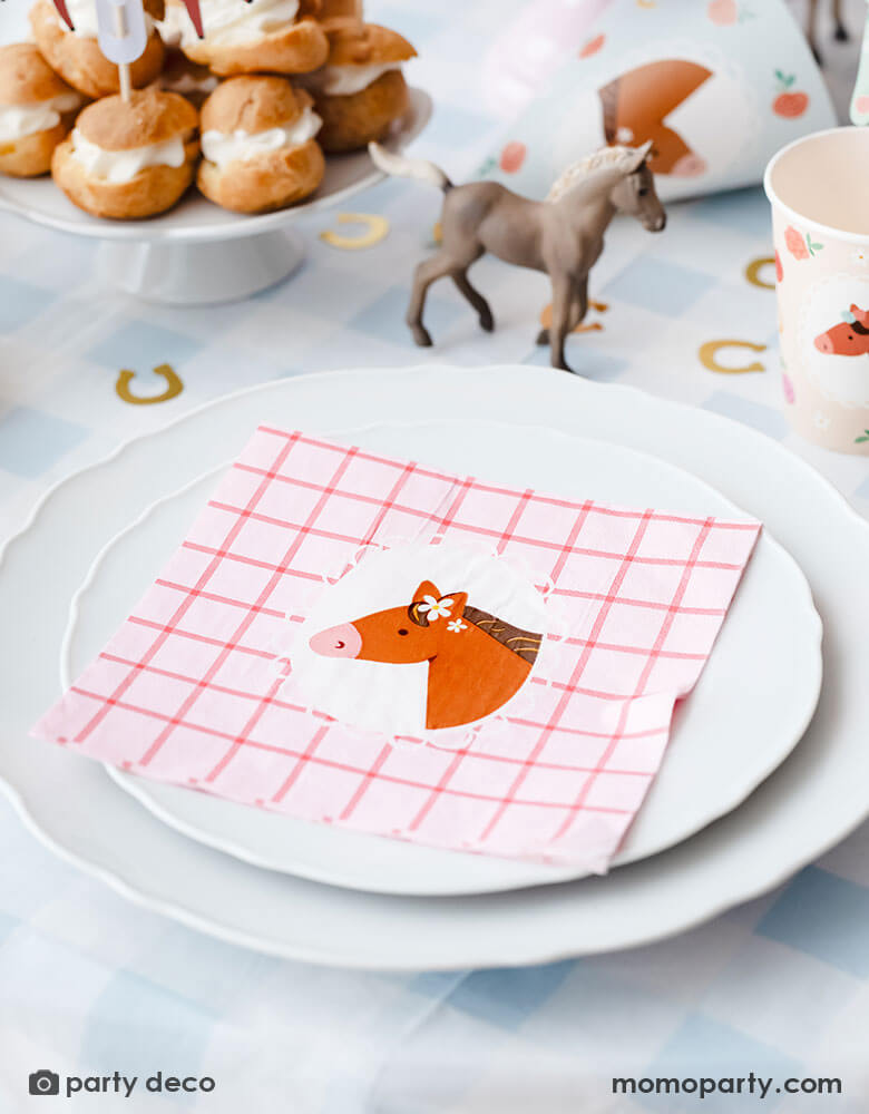 A pony horse themed birthday party table place setting featuring Momopartys's floral pony themed party supplies including the blue gingham tablecloth, large floral pony pink gingham napkins, party cups, and gold horseshoe shaped confetti around. On the table you can see some cream puff stacked in the back as dessert and a small horse figurine as the centerpiece - a great inspiration for kid's pony or horse themed party decoration.