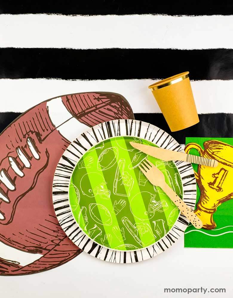 A modern yet festive football party tablescape featuring football themed tableware from Momopartys including the football shaped paper placemat, the touchdown dessert plates, the trophy shaped napkins on a black striped table runner, making this a perfect inpo for a kid's football themed birthday party or a Superbowl party table decoration. 