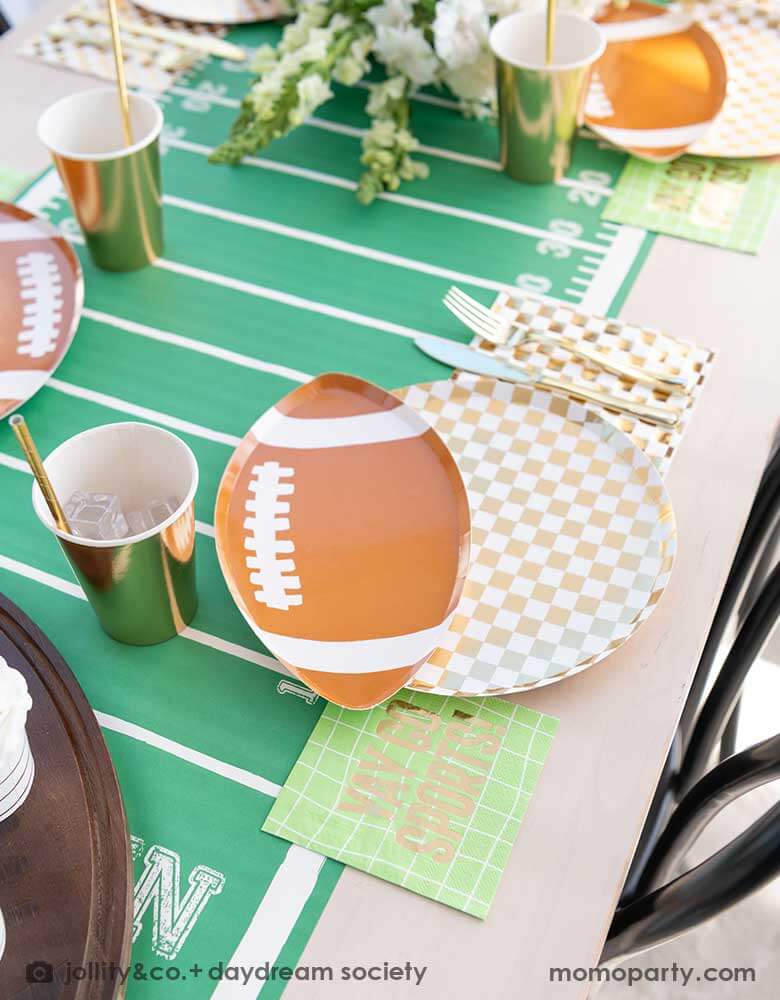 A modern football party table filled with Momopartys's football themed party tableware by Daydream Society including the die-cut football shaped plate, gold and white checkered dinner plate with green and coordinating napkins and gold party cups. In the center of the table there's a football field table runner and a beautiful flower bouquet, all makes great inspirations for football themed celebrations, a Super Bowl watching party or a Sunday football viewing party. 