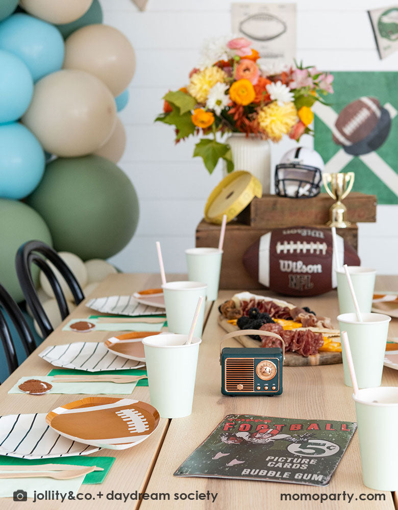 A modern kid's football birthday party table filled with Momopartys's football themed party tableware by Daydream Society including the die-cut football shaped plates, black striped plates, green and mint napkins and party cups. In the center of the table there's a cheese charcuterie board and some decorations like a football, a trophy and a football helmet as the centerpiece. On the wall there are some old school football posters and color matching balloons to add some festivities to the celebration!