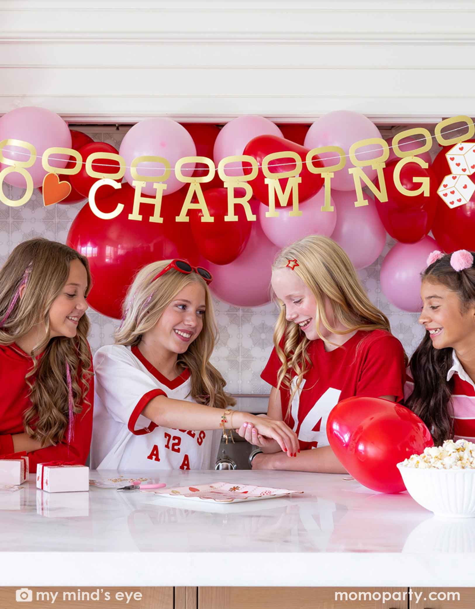 Four girls sitting at a Valentine's Day party table filled with heart themed party supplies from Momopartys including plates, napkins, and a cake. In the back there's a heart-themed balloon arch in red and pink and a gold foil party banner spells "So Charming". 