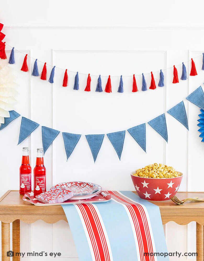 A wall decorated with Momopartys's patriotic party decorations and tableware by My Mind's Eye. Including a red, white and blue tassel banner, a blue flag banner with party paper fans. On the table there's the Hampton red and blue striped paper table runner, a red star bamboo tray with Hampton floral plates and napkins, next to two red soda bottles. On the right there's a big red star bamboo bowl with popcorn in it. Making this a great inspo for an elegant 4th of July home decor.
