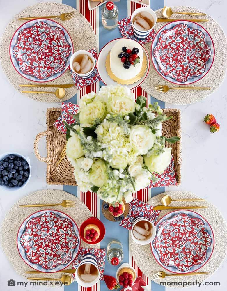 An elegant patriotic American themed party table features Momopartys's Hamptons Party Collection by My Mind's Eye including 10" Chambray Gingham Paper Plates, 10" Hampton Floral Wave Plates and napkins on white round placemats. In the middle of the table there are blue and red cakes, treats and drinks, along with a white floral bouquet as the centerpiece in the center on the Hampton striped paper runner, making this a perfect tablescape inspo for a 4th of July party or Memorial Day celebration.