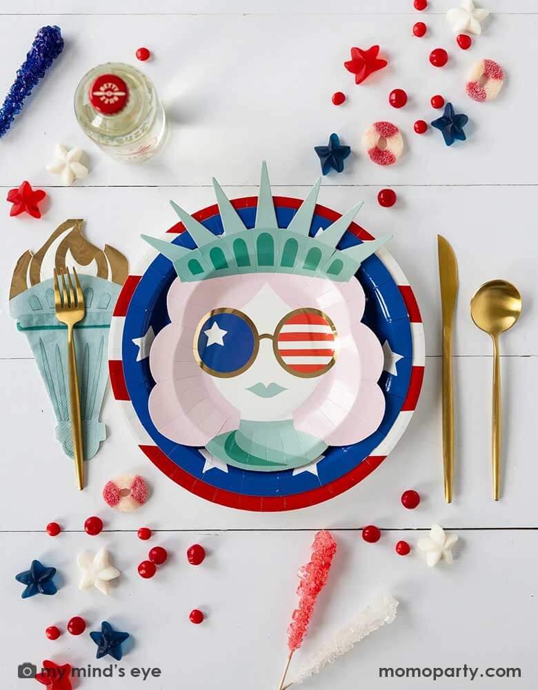 A modern and fun placeset featuring Momopartys's Lady Liberty Party Collection by My Mind's Eye including the 10" red striped round plate, 9" blue star round plate, lady liberty shaped plate, torch shaped dinner napkins and. Around them are various candies and treats in patriotic themed in red white and blue. A perfect party inspo for a modern Fourth of July celebration with friends and family.