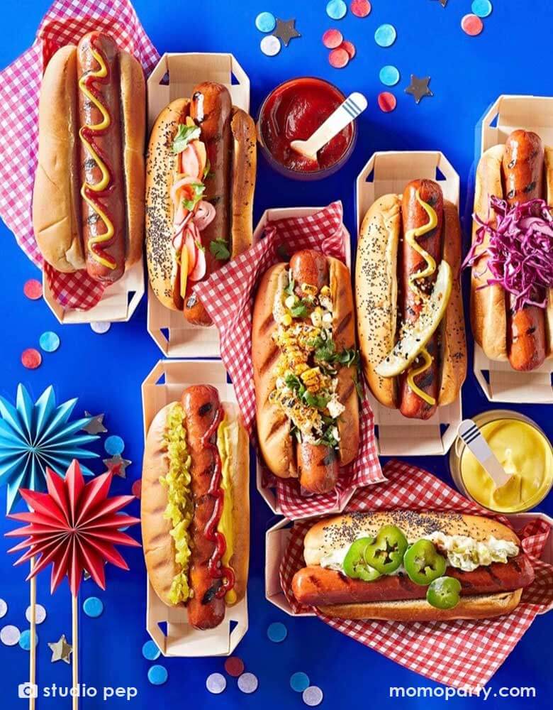 A table filled with different flavor of hotdogs with various condiment in hotdog paper containers adorned with red checkered paper, on the blue party table is Momopartys's patriotic confetti in red, white and blue spread out and some mini paper fan toppers in blue and red, making this a perfect inspo for a festive fourth of July party table and food ideas.