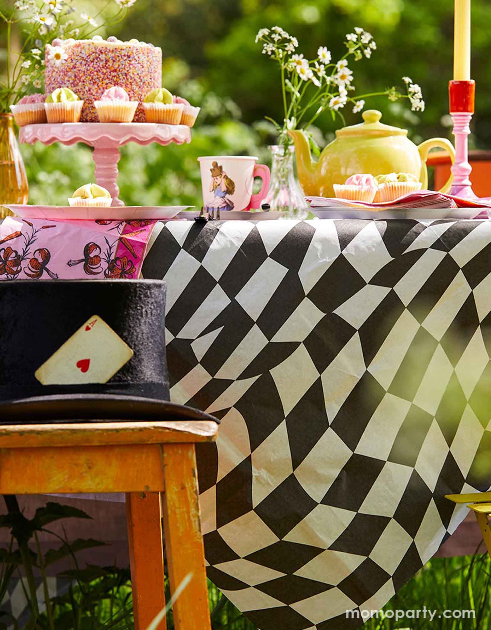 An Alice Wonderland themed garden tea party table set up featuring whimsical party supplies including plates, party cups and napkins on the black and white check table cover from Momopartys. With a birthday cake, some cupcakes a tea pot set with long candles and a magician's hat on the chair, a magical journey is about to start!