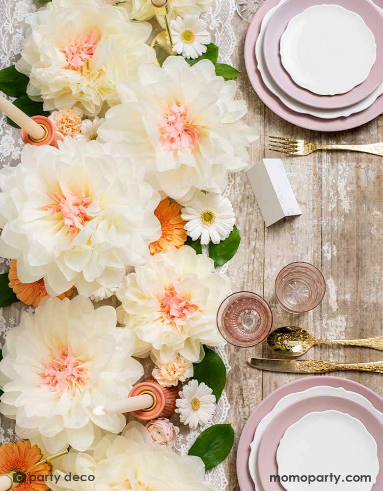 An elegant table set featuring layers of pink plates stacked and elegant gold utensils. For the centerpiece is Momopartys's cream flower pom pom decorations and blush candles. Making this a great inspiration for a rustic dinner table setting idea or wedding tablescape ideas.
