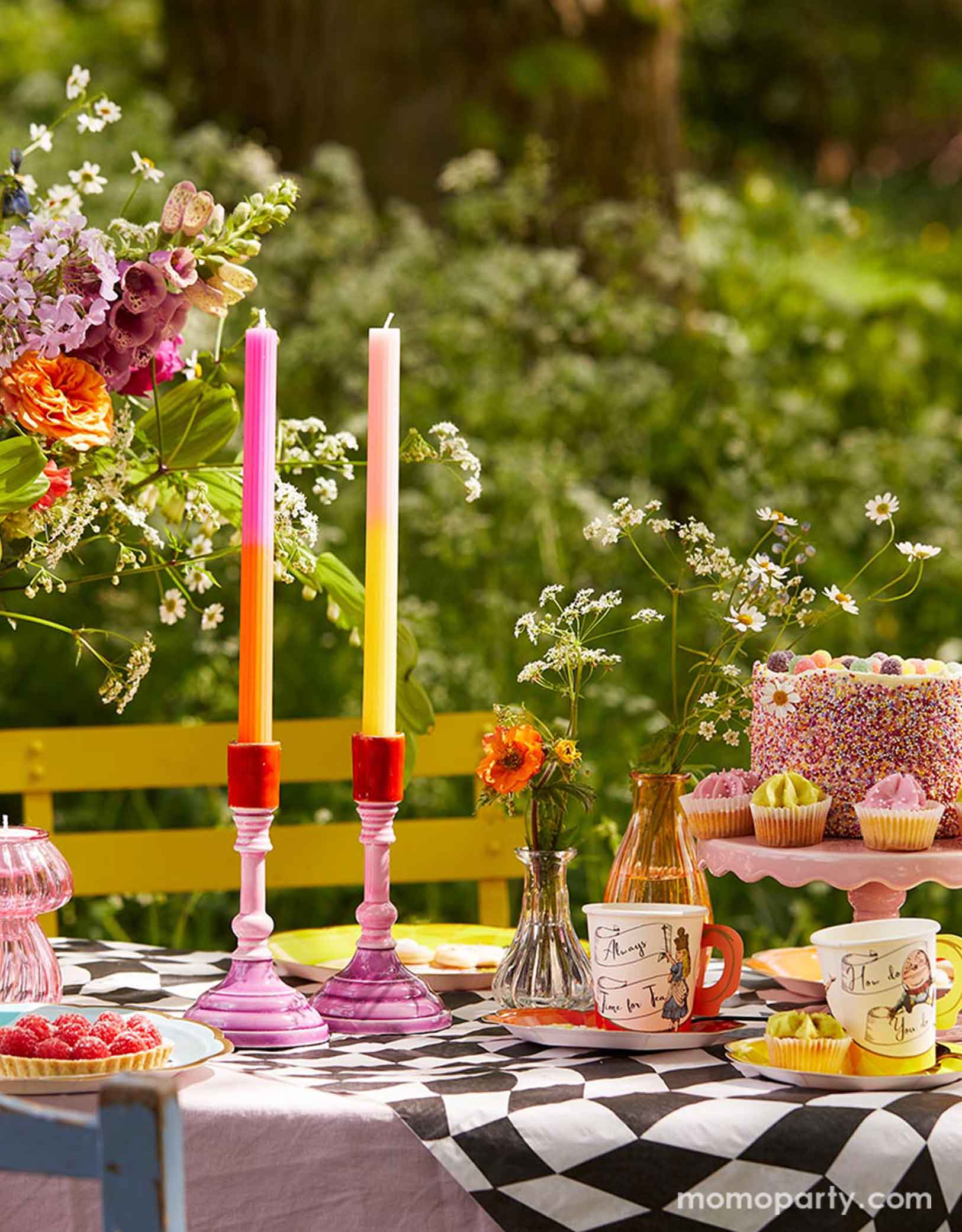 An Alice Wonderland themed garden tea party table set up featuring whimsical party supplies including plates, party cups and napkins on the black and white check table cover from Momopartys. With some flowers in glass vases, a birthday cake, some cupcakes, desserts, long candles on the table, a magical journey is about to start!
