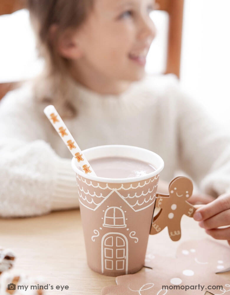 A girl in a white sweater holding Momopartys's 12 oz Gingerbread Handled Paper Cup with a festive gingerbread man handle in a gingerbread house themed Holiday party. With gingerbread man shaped napkins on the table this creates a cozy yet festive vibe for a whimsical Holiday party for kids.