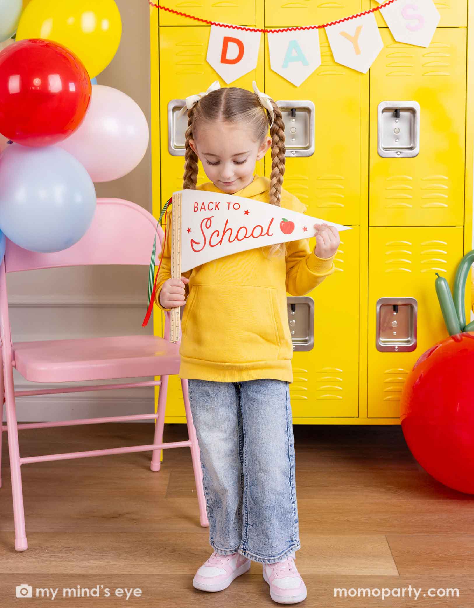 A girl in yellow shirt standing in front of a first day of school celebration decoration set up featuring various school themed party decors from Momopartys including School Days felt party banner and a festive balloon garland. She's holding the ruler back to school felt party pennant and looking at it. There's a yellow locker wall behind her and a pink folding chair. Making this a cute party decoration set up for kid's first day of school celebration.