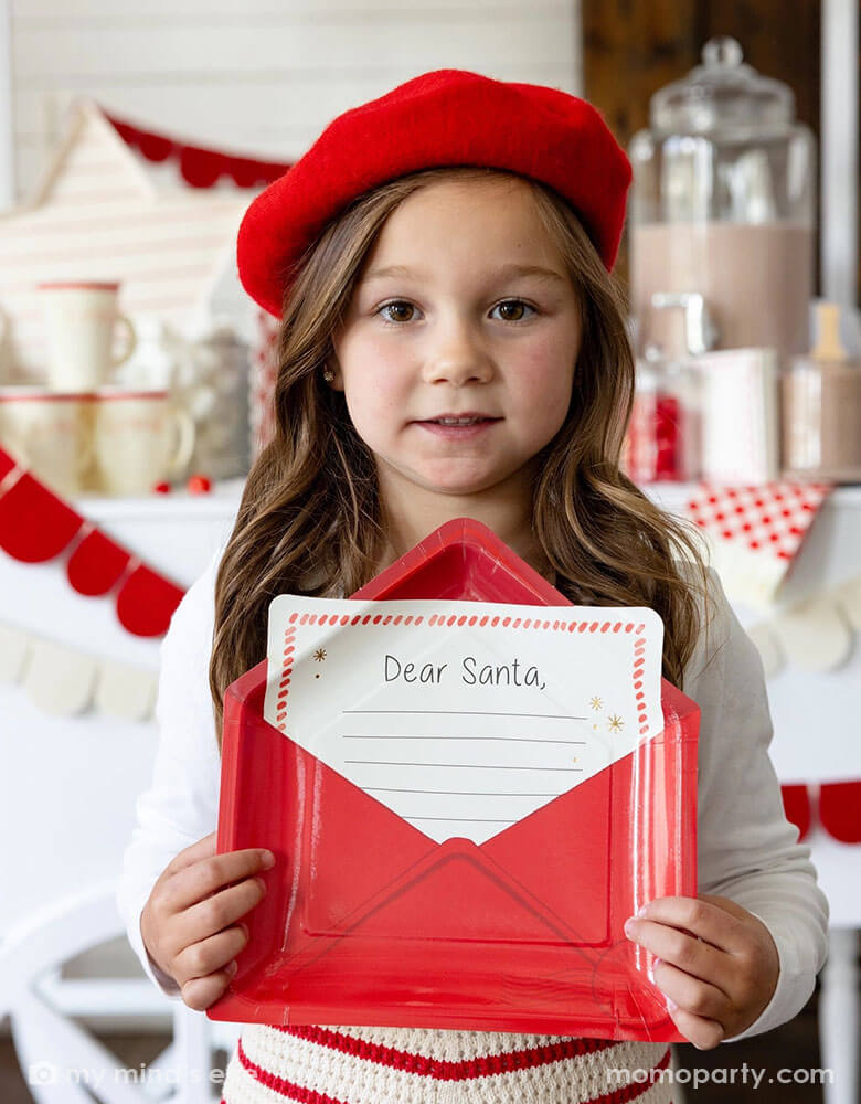 A school aged girl dressed in her Holiday outfit with a red beret holding Momopartys's Letter to Santa envelope shaped plate in front of a hot chocolate station of a table filled with Momopartys's Dear Santa party supplies including a red felt party garland, Santa shaped party cups, red checkered party plates and napkins next to hot cocoa and toppings, making this a cozy yet whimsical set up for a festive Holiday celebration!