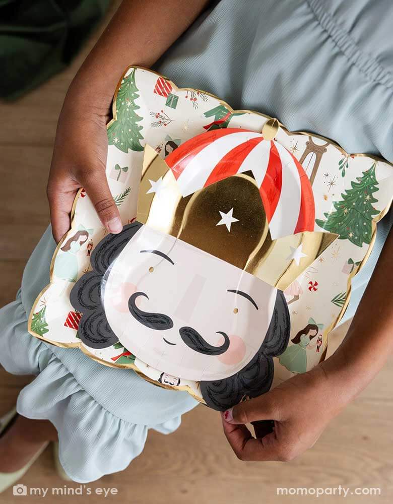 An overhead shot of a girl in her Holiday dress holding Momopartys's 10" Nutcracker pattern plate and Nutcracker head shaped plate in a playful design. 