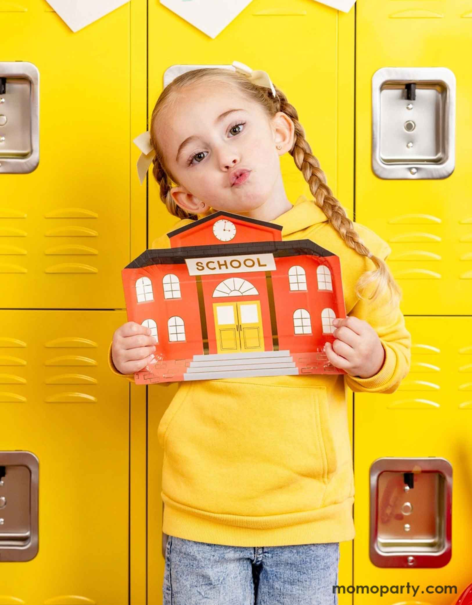A school aged girl with two braids holding Momopartys's 10.25" x 8" School House Shaped Paper Plate by My Mind's Eye. She's wearing a pencil inspired yellow sweatshirt standing in front of an array of vintage inspired yellow lockers at school.