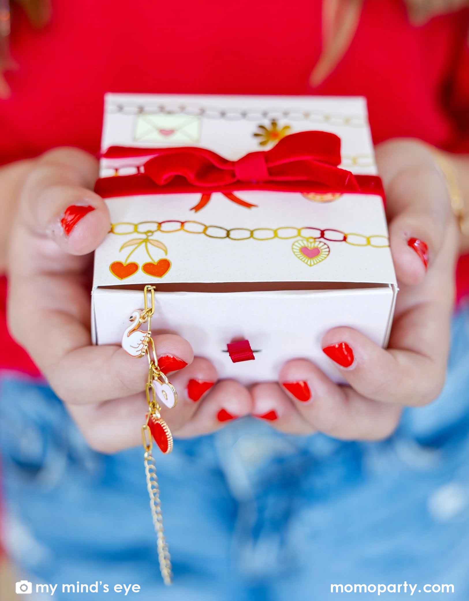 A girl with red nail polish is holding Momopartys's Valentine's Day Charm Bracelet Kit gift box with red ribbon and heart designs, accompanied by a gold bracelet.