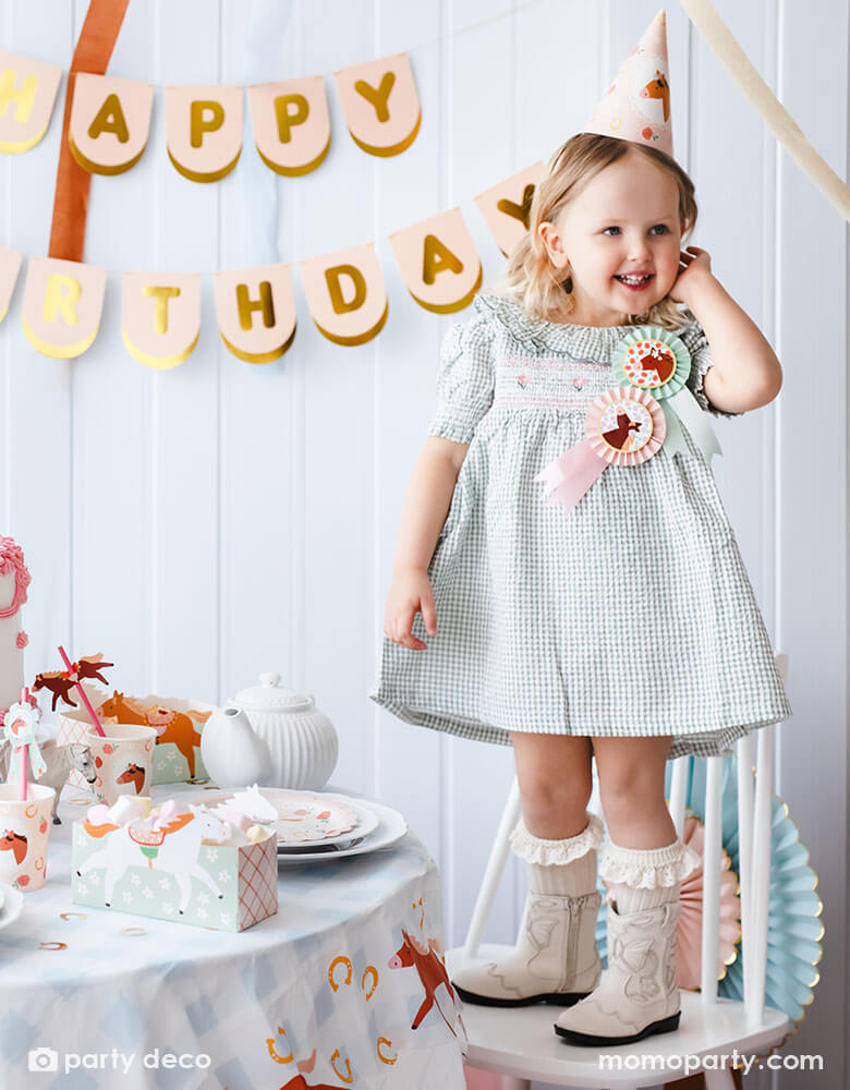 A preschool girl with a birthday party hat in a gingham dress and Momopartys's horse rosette ribbons on her chest, standing next to a party table featuring Momopartys's floral pony themed party supplies including floral pony party cups, plates, cupcake toppers, napkins treat boxes and paper straws, along with the blush happy birthday party banner hung on the wall - making this a perfect inspo for a girl's horse themed birthday party.