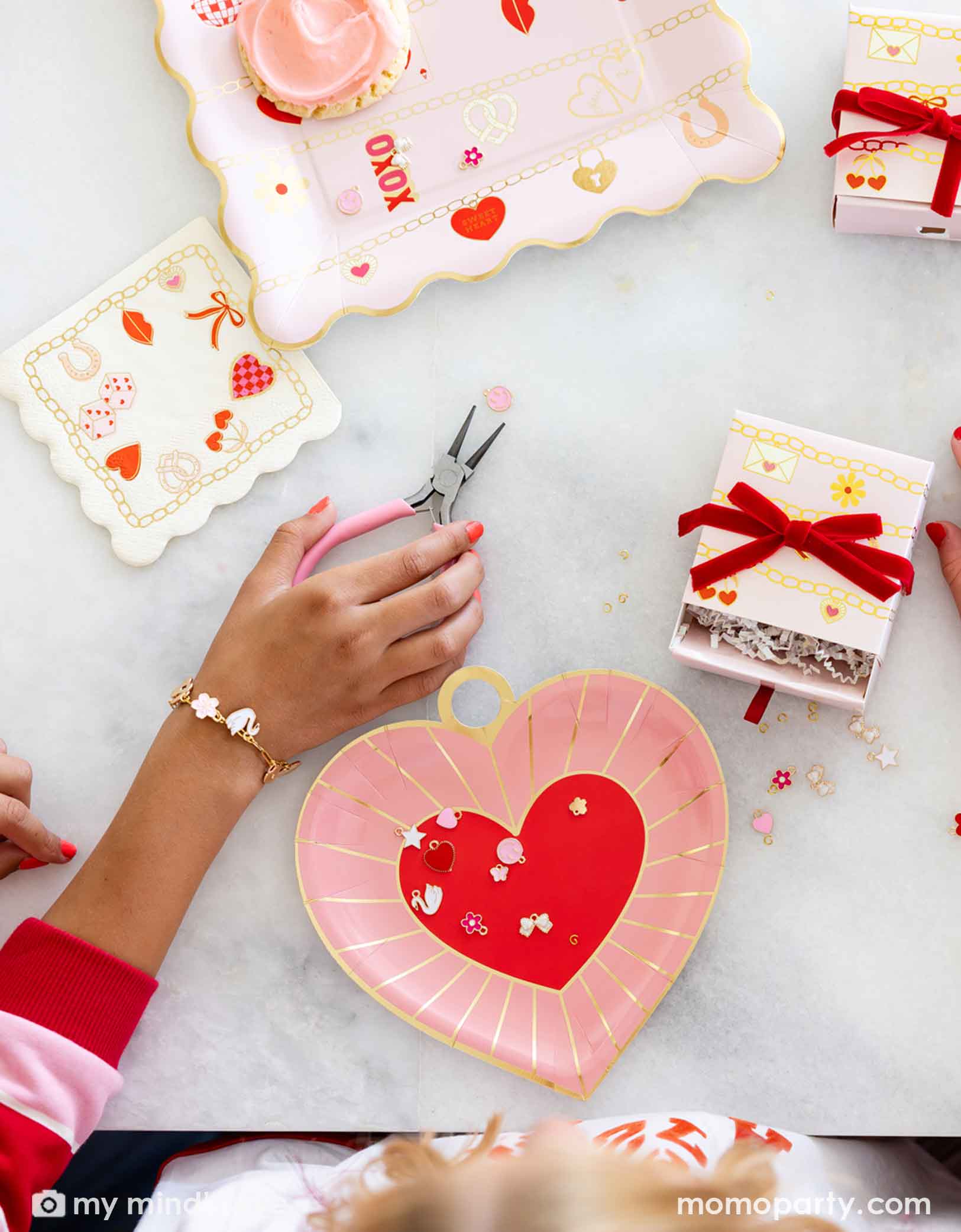 Children's hands with pink nail polish working on their charm bracelets with pink pliers, surrounded by small charms, gift boxes and Momopartys's Valentine's Day charm matching party tablewares including heart shaped plates and napkins on the table. 