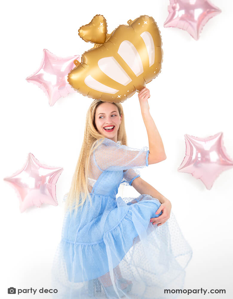 A blonde lady in a Cinderella-like light blue tutu dress holding Momopartys's 20" x 17" princess crown shaped foil balloon by Party Deco. In the back there are four pink star shaped foil balloons as the background, making this a good inspiration for a princess themed party decoration. 