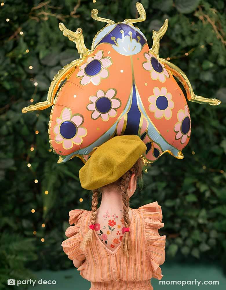 A little girl holding Momopartys's 34 x 30 inches ladybird shaped foil balloon by Party Deco. With coral color with small flower pattern with gold legs on it. She's dressed in her spring outfit with a mustard beret hat standing in a backyard with a wall filled of greenery.