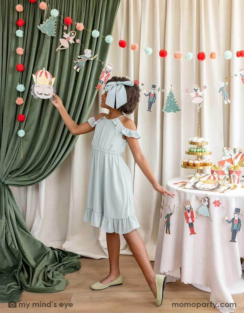 A school aged girl doing a ballet dance pose holding Momopartys's 10" Nutcracker head shaped party plate in the air, standing next to a party table filled with Holiday treats and Nutcracker themed party supplies including plates, cups, napkins and party garland. In her back there's a stage curtain in satin green and beige which is decorated with Momopartys's Nutcracker garland with pom-poms which adds a festive touch to the Holiday decoration.
