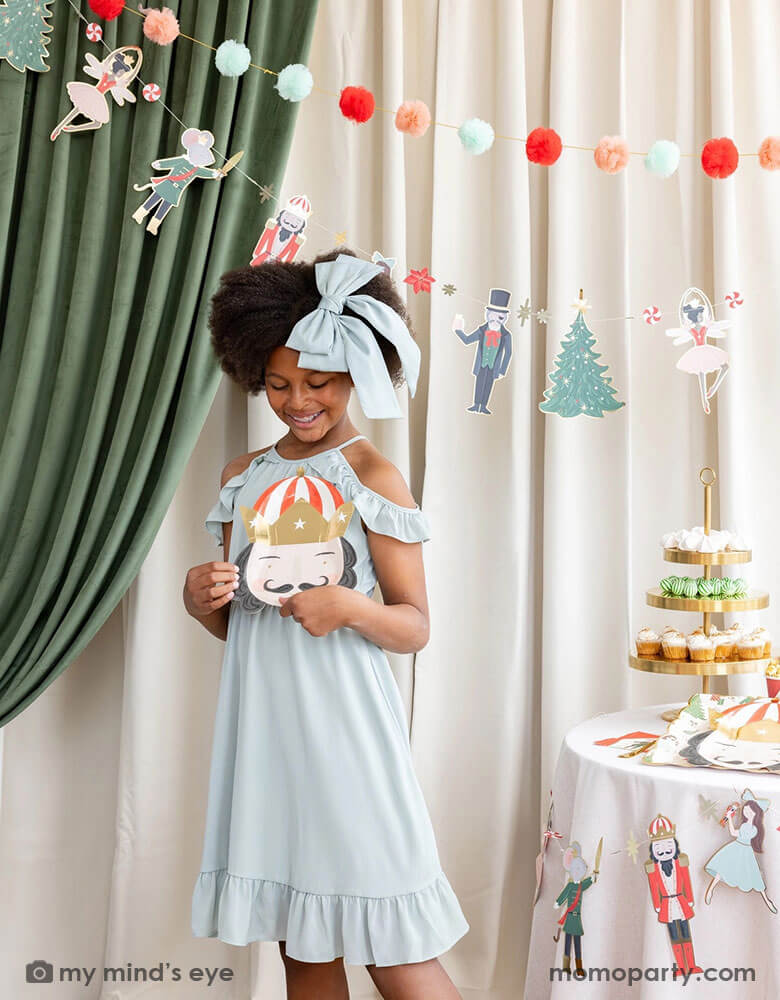 A school aged girl holding Momopartys's 10" Nutcracker head shaped party plate standing next to a party table filled with Holiday treats and Nutcracker themed party supplies. In her back there's a stage curtain in satin green and beige which is decorated with Momopartys's Nutcracker garland with pom-poms which adds a festive touch to the Holiday decoration.