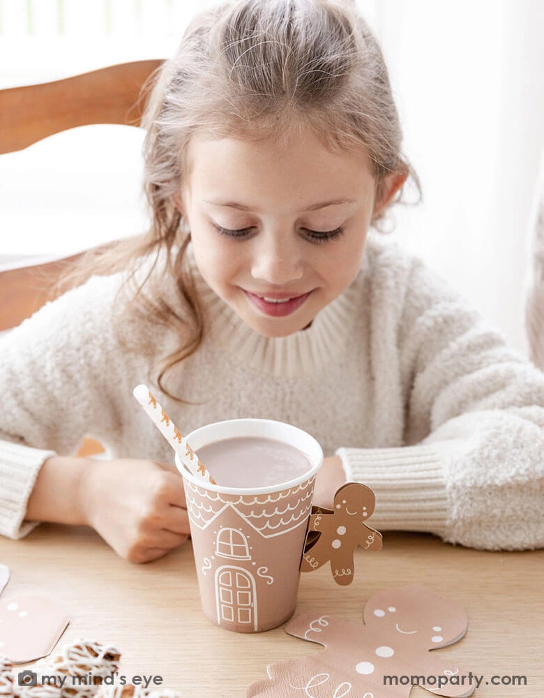 A girl in a white sweater looking into Momopartys's 12 oz Gingerbread Handled Paper Cup with a festive gingerbread man handle. There's hot chocolate in the cup with a gingerbread man straw, in a gingerbread house themed Holiday party. With gingerbread man shaped napkins and Holiday treats on the table this creates a cozy yet festive vibe for a whimsical Holiday party for kids.