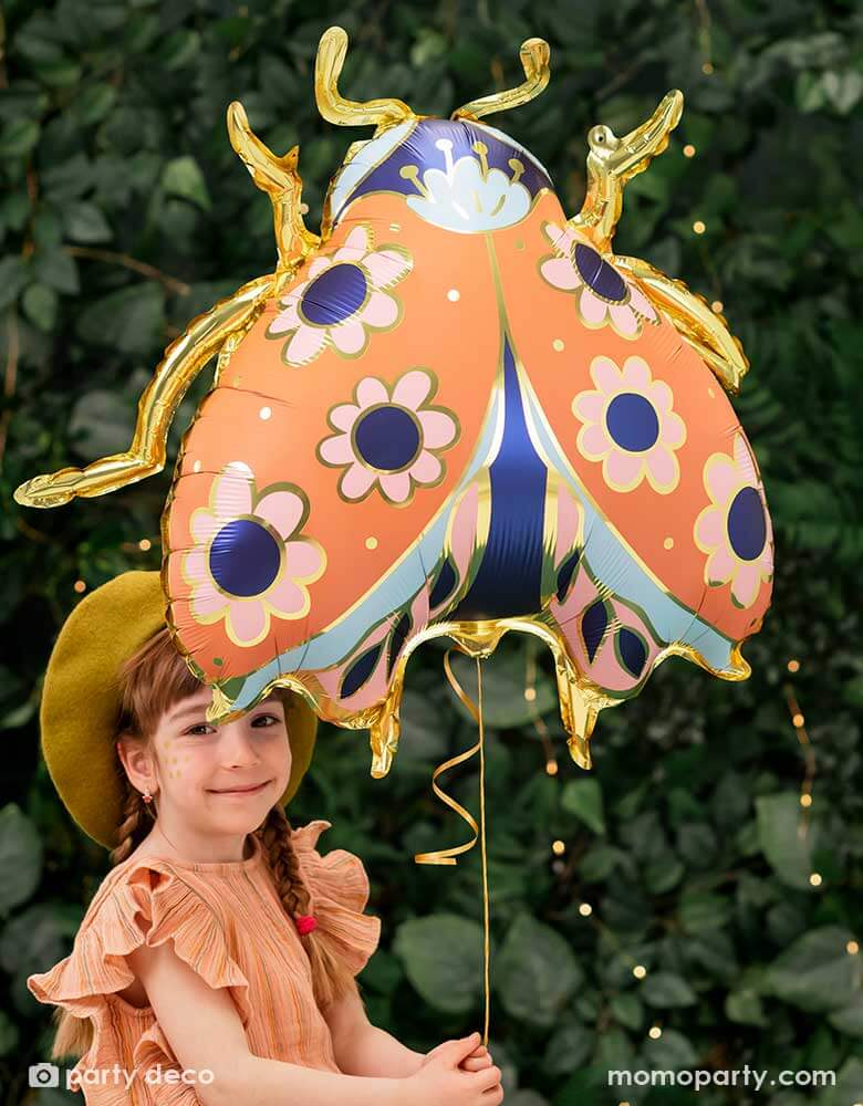 A little girl holding Momopartys's 34 x 30 inches ladybird shaped foil balloon by Party Deco. With coral color with small flower pattern with gold legs on it. She's dressed in her spring outfit with a mustard beret hat standing in a backyard with a wall filled of greenery.