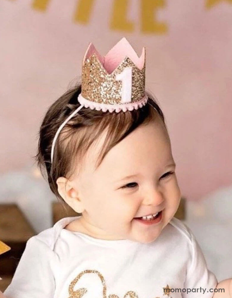 A one-year girl in her pale pink outfits wears Momopartys's 3" first birthday pink glittered birthday party crown. She has a sweet smile and in her back there's a pink party backdrop with gold happy birthday banner in the back.