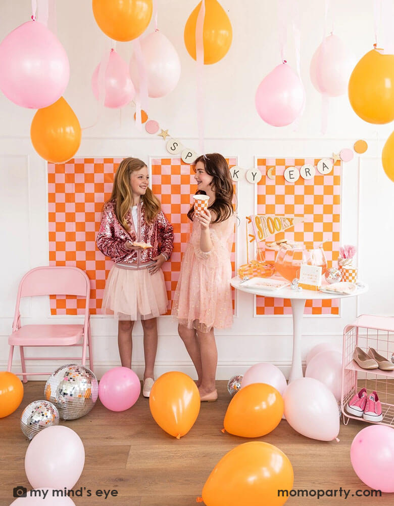Two middle school girls in their pink outfits chatting with each other in a groovy themed Halloween party decorated with party supplies and decorations in orange and pink. The wall is decorated with orange and pink checkered patterned posters and a Taylor Swift friendship bracelet inspired banner says "Spooky Era." The room is filled with balloons in orange and pink and there's a table full of Halloween party supplies including trendy plates, cups, napkins, pennants from Momopartys.