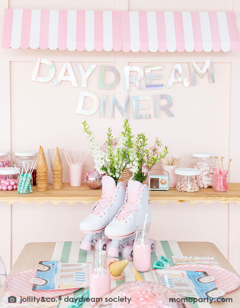 An adorable kid's ice cream themed birthday party tablescape featuring Momopartys's ice cream truck shaped plates, the pink checkered plates, the ice cream icon napkins on a mint striped table runner. On the table there's a roller skate and pink disc balls as centerpiece. Behind the table there's a sundae station with sprinkle toppings, ice cream cones, adorned with a retro pink striped awning as a small ice cream shop, making this a cute and festive ice cream party set up for kids birthday celebration.