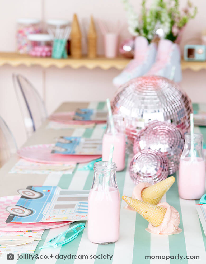 An adorable kid's ice cream themed birthday party tablescape featuring Momopartys's ice cream truck shaped plates, the pink checkered plates, the ice cream icon napkins on a mint striped table runner. On the table there's a roller skate, pink disc balls and ice cream cone decorations as the centerpiece. Behind the table there's a sundae station with sprinkle toppings, ice cream cones, making this a cute and festive ice cream party set up for kids birthday celebration.