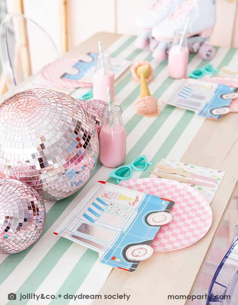 An adorable kid's ice cream themed birthday party tablescape featuring Momopartys's ice cream truck shaped plate, the pink checkered plate, the ice cream icon napkin and pink wooden cutlery on a mint striped table runner. Around the placeset there are some summer inspired decoration like a pair of mint sunglasses, pink disco balls, a pair of pink roller skates, some milk bottles with gum ball pink drinks, and some ice cream cone decorations, making this a cute, fun and festive ice cream social.