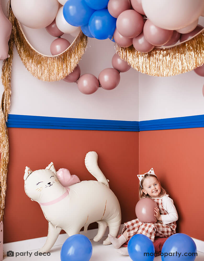 A girl dressed like a cat sitting next to the kitty cat shaped foil balloon from Momopartys. Around them the room is decorated with festive balloons in different shades of pink and royal blue witch matches the colors of the walls. Along with gold tinsel fringe garlands, it creates a fun and festive atmosphere for kid's pet or cat themed birthday celebration.