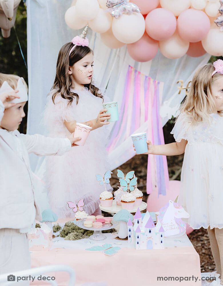 A few kids dressed in beautiful dresses in a dreamy fairytale themed birthday party table featuring Momopartys's unicorn princess fairy themed party including butterfly cupcake toppers, castle centerpiece and butterfly shaped napkins. The kids are holding Momopartys's 7.4 oz pastel bow paper cups in 3 different colors. In the back there's a pastel balloon cloud adorned with butterfly foil balloons and a white curtain, making this a whimsical set up for kid's fairytale birthday celebration. 