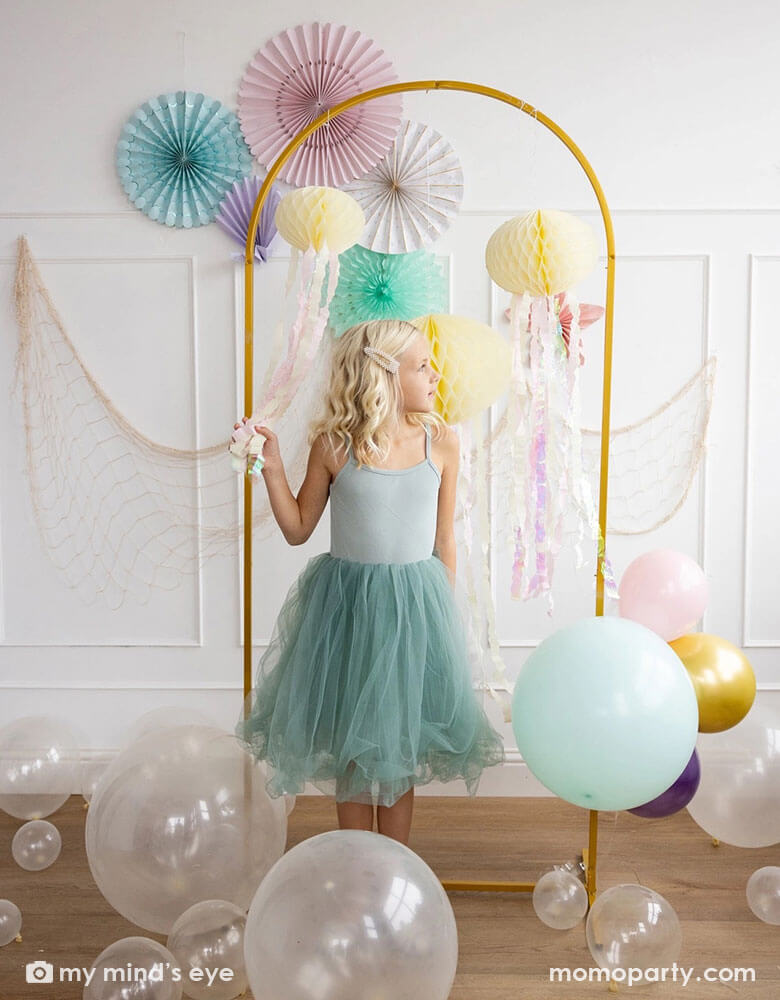 A young girl in her teal tutu dress standing in front of a white wall decorated with mermaid inspired party decorations including Momopartys's under the sea paper fans in pastel colors of aqua, pink, lilac, teal, cream and peach. Next to the paper fans hung a fishing net draping over a golden arch structure adorned with honeycomb jelly fish decorations by My Mind's Eye. On the floor there are some clear latex balloons as bubbles, making this an inspo for an enchanting kid's mermaid birthday party set up.