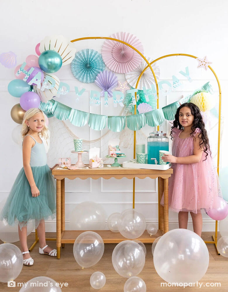 Two school-aged girls in an enchanting kid's mermaid birthday party set up featuring mermaid inspired decorations and balloons from Momopartys including the Under the Sea paper fan set in pastel colors, Mermaid Happy Birthday Banner Set and shell shaped paper decorations by My Mind's Eye on the wall. The girls are standing by a party table with mermaid inspired party supplies including cups, plates, napkins and treat boxes. On the floor are some clear latex balloons creating a whimsy water bubble scene.