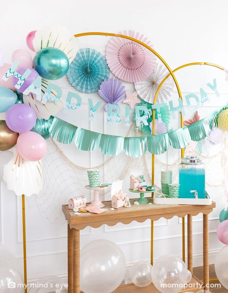 An enchanting mermaid party set up features Momopartys's mermaid decorations and and supplies by My Mind's Eye including including the Under the Sea paper fan set in pastel colors of light pink, aqua, teal, lilac and cream, Mermaid Happy Birthday Banner Set with aqua foil fringe banner and shell shaped paper decorations on the wall. On the table there are various mermaid themed tableware including cups, napkins, plates and treat boxes. On the floor are clear latex balloons creating a whimsy bubble scene.