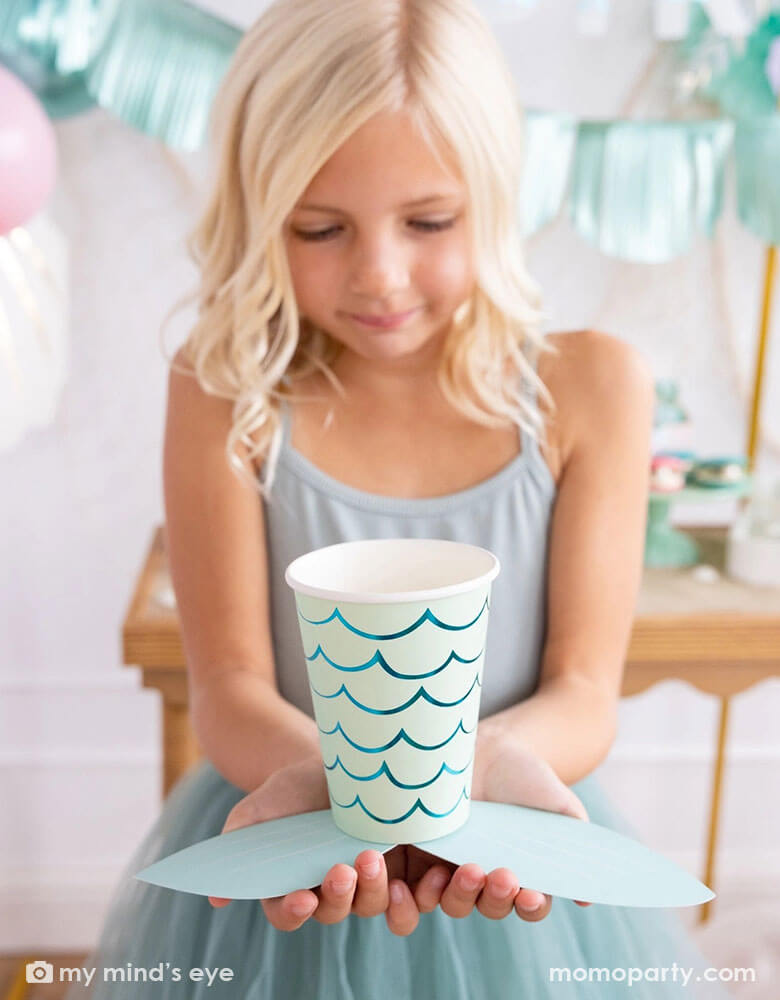 A school-aged girl in a teal tutu dress holding Momopartys's mermaid tail paper cups by My Mind's Eye. In her back you can see some mermaid party decorations on the wall behind a party table featuring teal and aqua themed party supplies for a under the sea/mermaid birthday celebration.