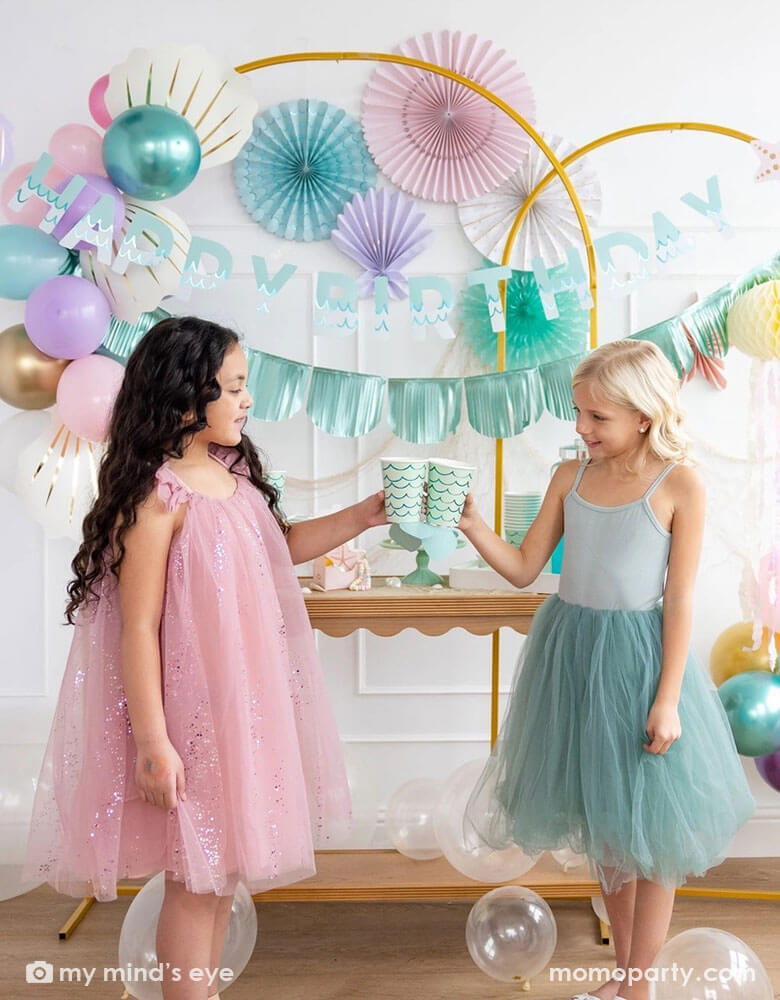 Two school-aged girls in their teal and pink tutu dresses celebrating a birthday in an enchanting kid's mermaid birthday party set up featuring mermaid decorations and balloons from Momopartys including the Under the Sea paper fan set in pastel colors of light pink, aqua, teal, lilac and cream, Mermaid Happy Birthday Banner Set with aqua foil fringe banner and shell shaped paper decorations by My Mind's Eye on the wall. The girls are raising their aqua mermaid tail party cups and cheering to each other.