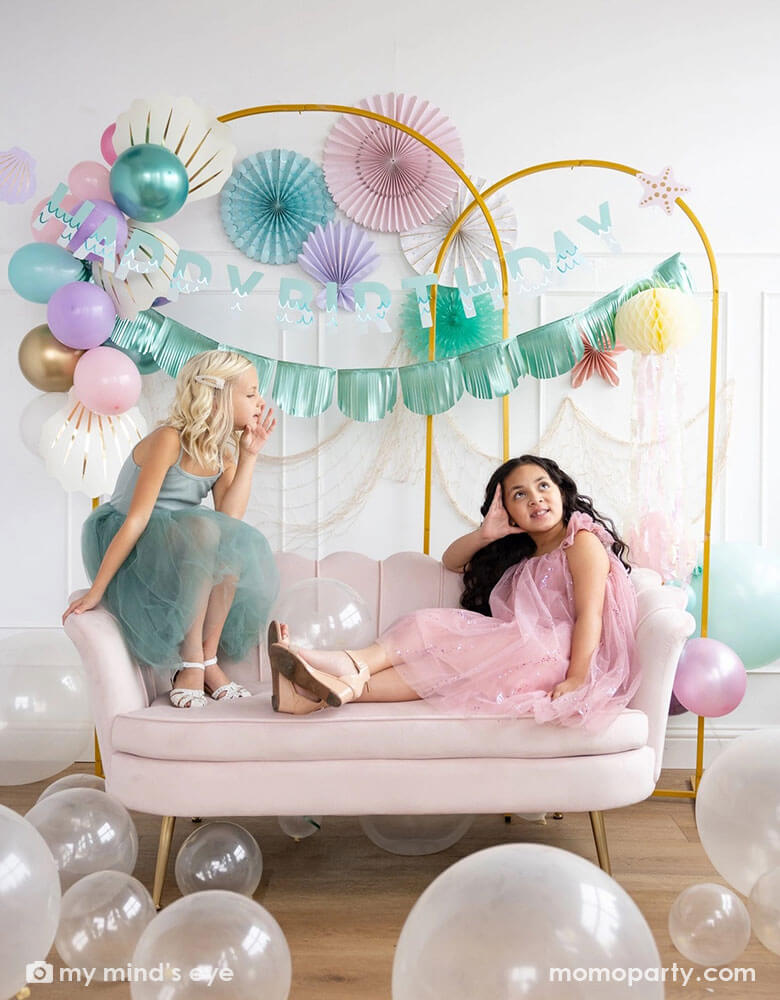 Two school-aged girls sitting on a shell shaped blush sofa in an enchanting kid's mermaid birthday party set up featuring mermaid inspired decorations and balloons from Momopartys including the Under the Sea paper fan set in pastel colors of light pink, aqua, teal, lilac and cream, Mermaid Happy Birthday Banner Set with aqua foil fringe banner and shell shaped paper decorations by My Mind's Eye on the wall. On the floor are some clear latex balloons in various sizes creating a whimsy water bubble scene.