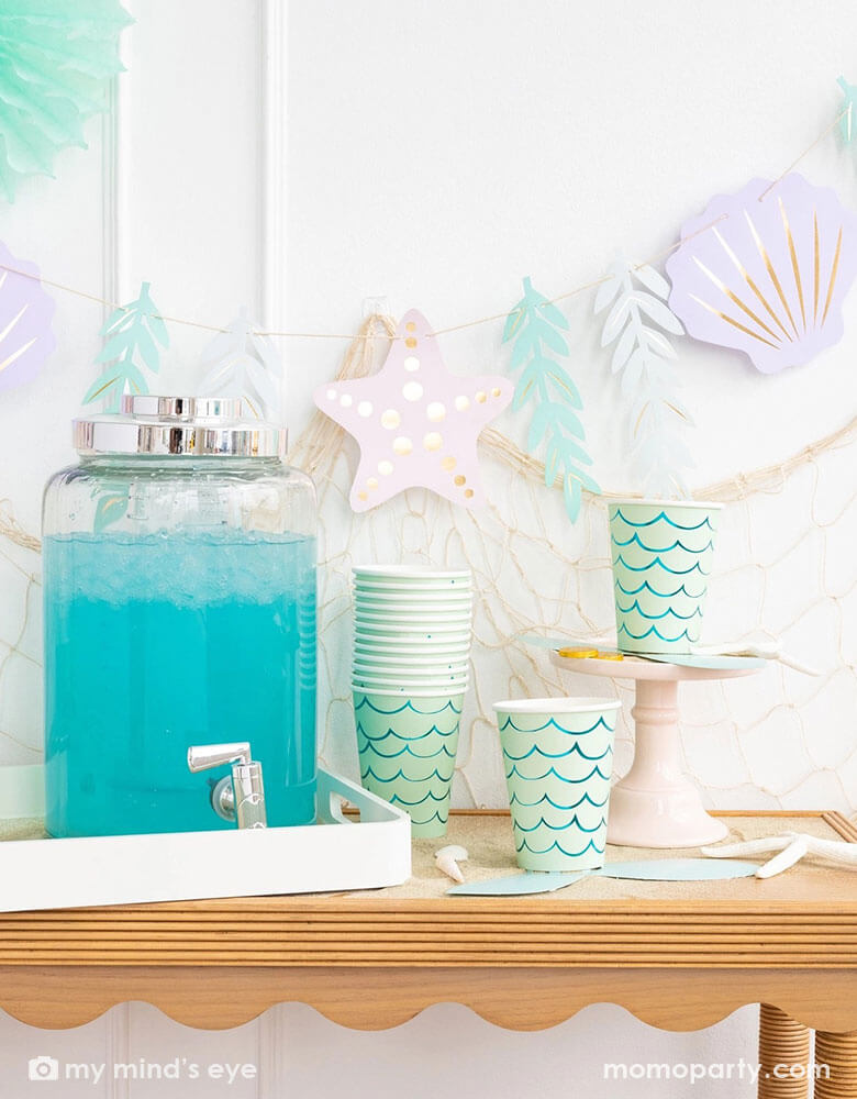 A mermaid party table features Momopartys's mermaid tail paper cups by My Mind's Eye. Next to the paper cups is a water dispenser filled with aqua colored drink. The table is decorated with shells, sea star and white sand decorations. In the back there's a fishing net hung on the wall which is adorned with under the sea paper party pennants features sea stars, sea grass, and sea shells in soft pastel colors, making this a perfect inspo for a kid's mermaid, under the sea party decorations.