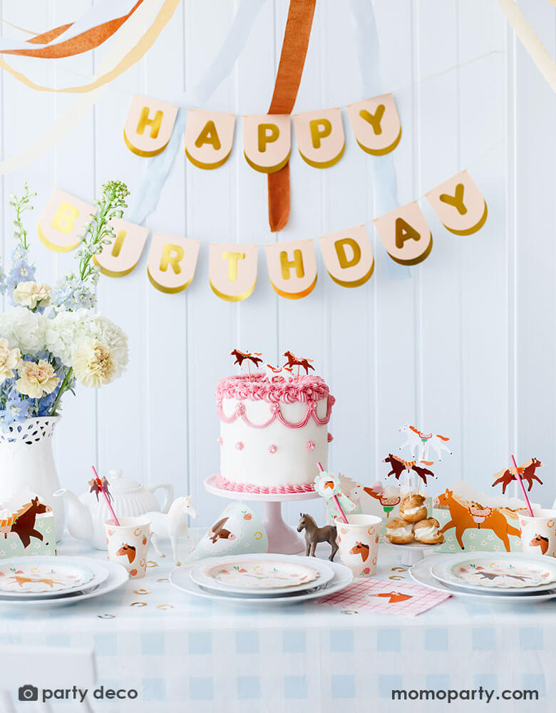 A kid's pony horse themed birthday party table decoration featuring Momopartys's floral pony themed party supplies including floral pony party cups, plates, cupcake toppers, napkins treat boxes and paper straws. In the middle of the table is a pink and white buttercream cake topped with pony shaped birthday candles, along with the blush happy birthday party banner hung on the wall and floral arrangement, this table setting makes a great inspo for a charming horse themed birthday celebration for girls.