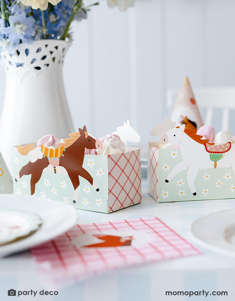 A horse/pony themed birthday party table features Momopartys's 6.7" x 4.3" x 2.95" floral pony treat boxes by Party Deco. Each snack box has some delicious treats in them including cookies and macarons. Behind the treat boxes is a beautiful flower arrangement and some party hats. In front of the snack boxes is a place setting features a pink gingham pony napkin -  a darling inspo for girl's pony themed party.