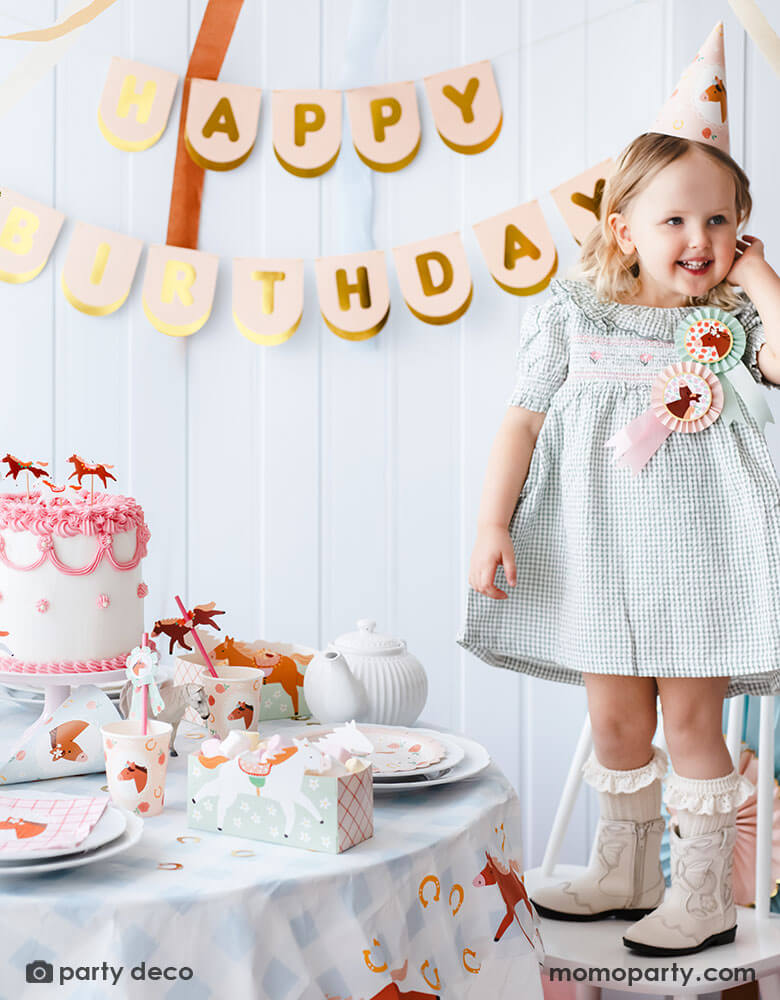 A preschool girl with a birthday party hat in a gingham dress and blush cowgirl boots standing next to a party table featuring Momopartys's floral pony themed party supplies including floral pony party cups, plates, cupcake toppers, napkins treat boxes and paper straws. In the middle of the table is a pink and white buttercream cake topped with pony shaped birthday candles, along with the blush happy birthday party banner hung on the wall - a perfect info for a girl's horse themed birthday party.